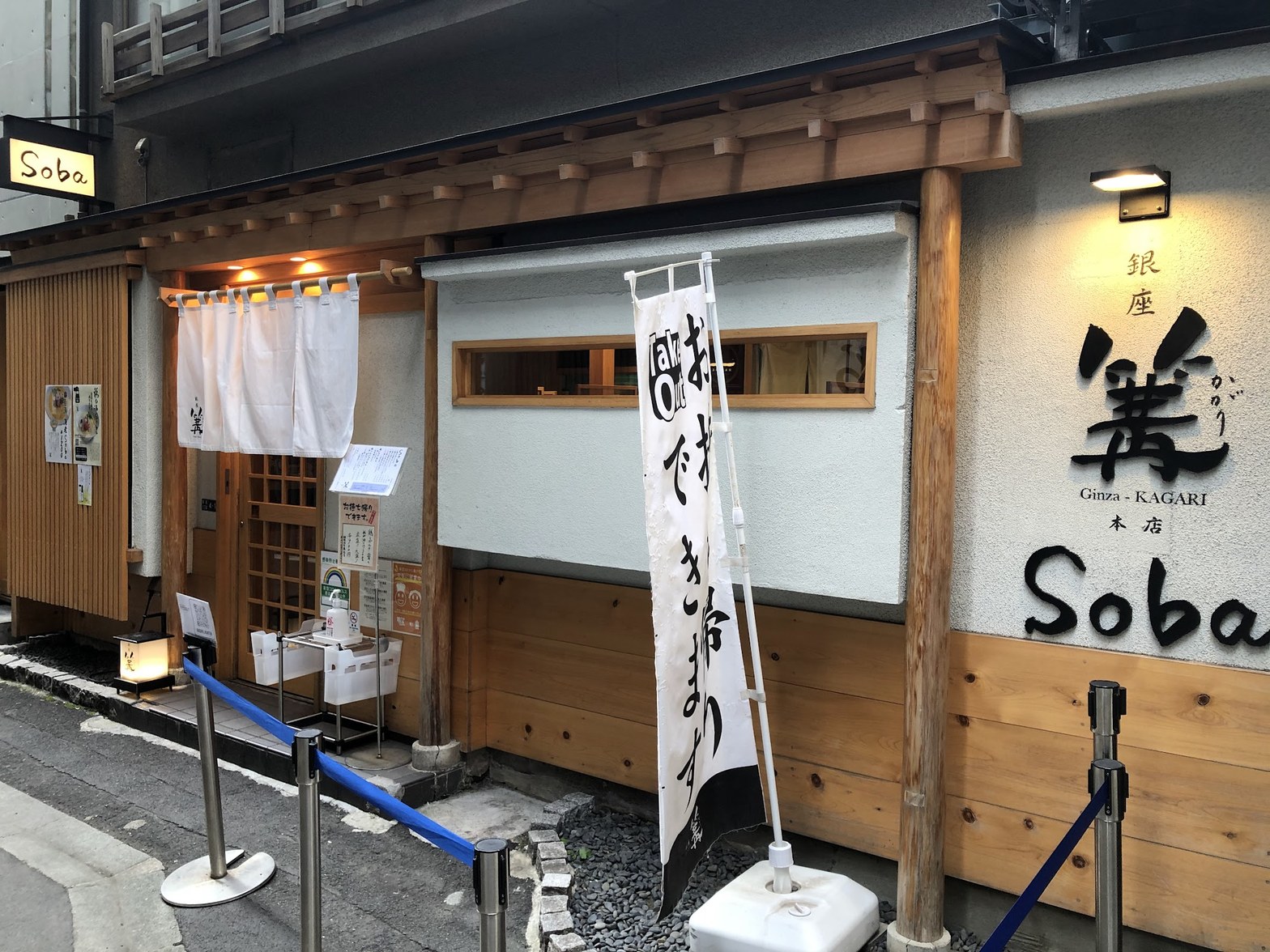 木製外壁と白のれん・「お待ちできます」ののぼり・青いパーティションが並ぶ篝本店の昼間外観。右壁面に大きな篝の筆文字サイン