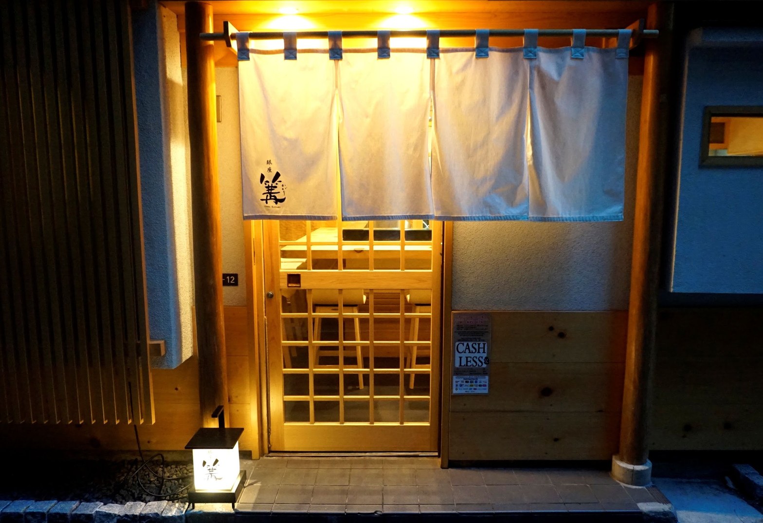 夜の篝本店入口。白のれんに「銀座 篝」の墨文字、木製格子扉の向こうに温かみのある照明と店内カウンターが透けて見える