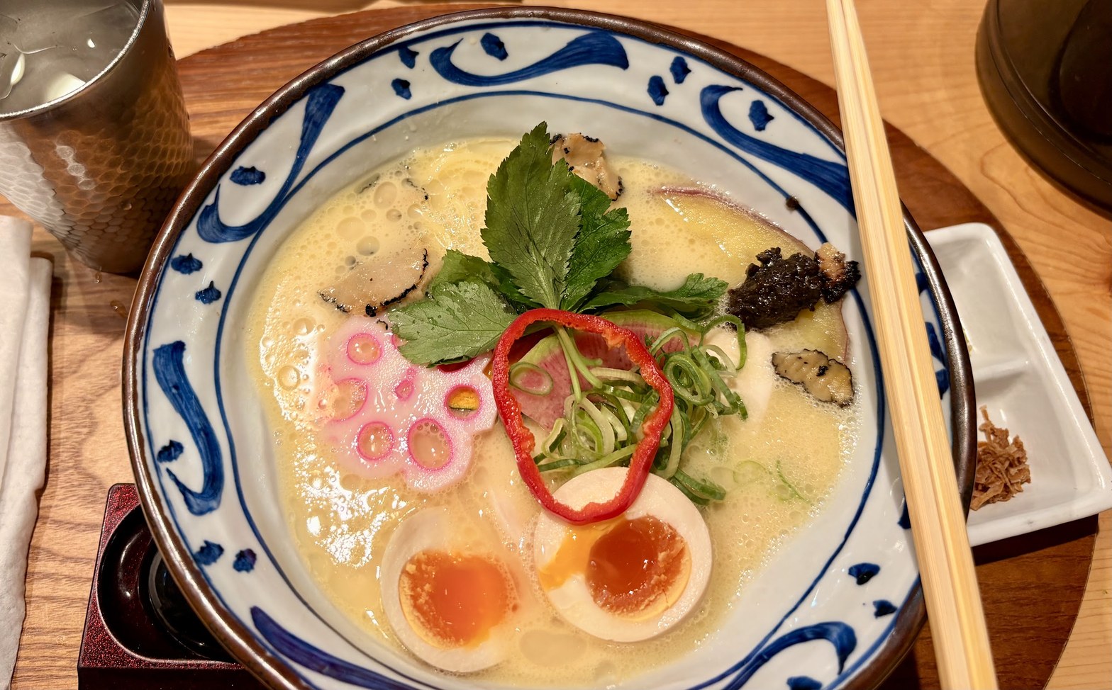 藍染め文様の丼に盛られた鶏白湯ラーメン。白濁したクリーミーなスープに味玉・赤パプリカ・三つ葉・トリュフ・なると・青ねぎが乗り、木製箸が添えられている