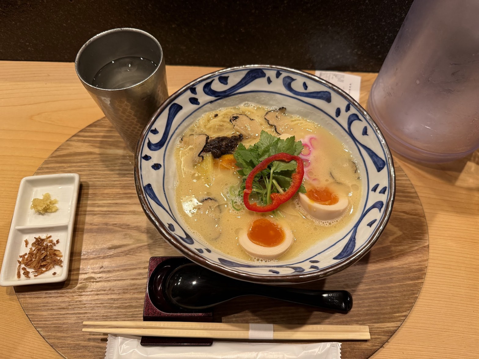丸いカウンターテーブルに置かれた藍染め文様の丼。白濁した鶏白湯スープに味玉・赤パプリカ・三つ葉・トリュフが乗り、黒レンゲと箸が添えられ、薬味小皿が横に並ぶ