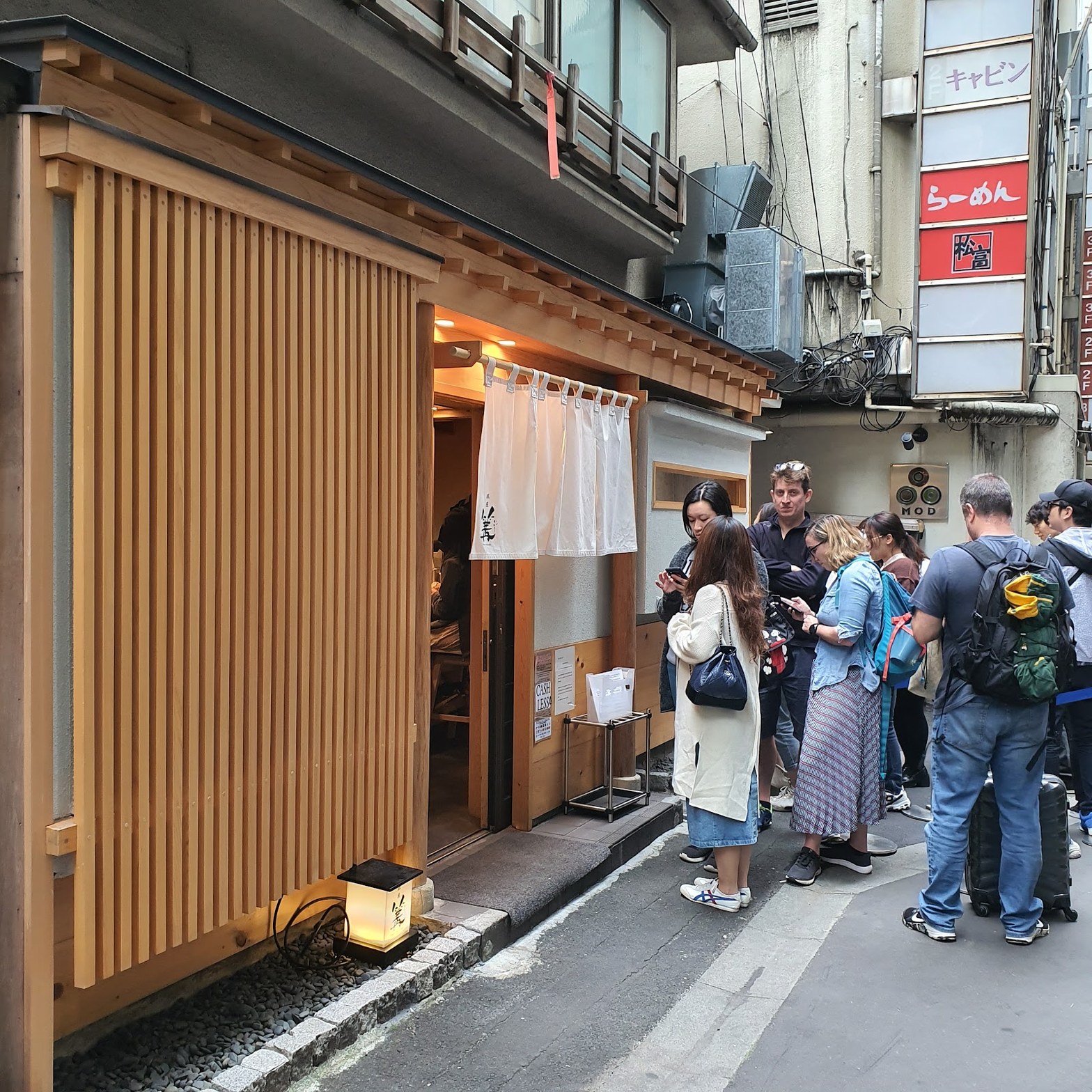 銀座裏路地の篝本店外観。木格子の壁と白のれんの入口前に外国人観光客を含む複数人が並んでいる昼間の光景